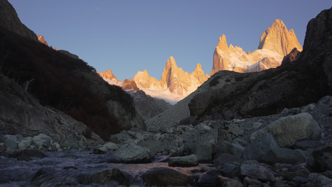 L'alba illumina il Fitz Roy e il Monte Saint Exupéry sopra un fiume in Patagonia, mostrando cime aspre e acqua che scorre