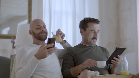 happy gay couple having breakfast in bed and using gadgets