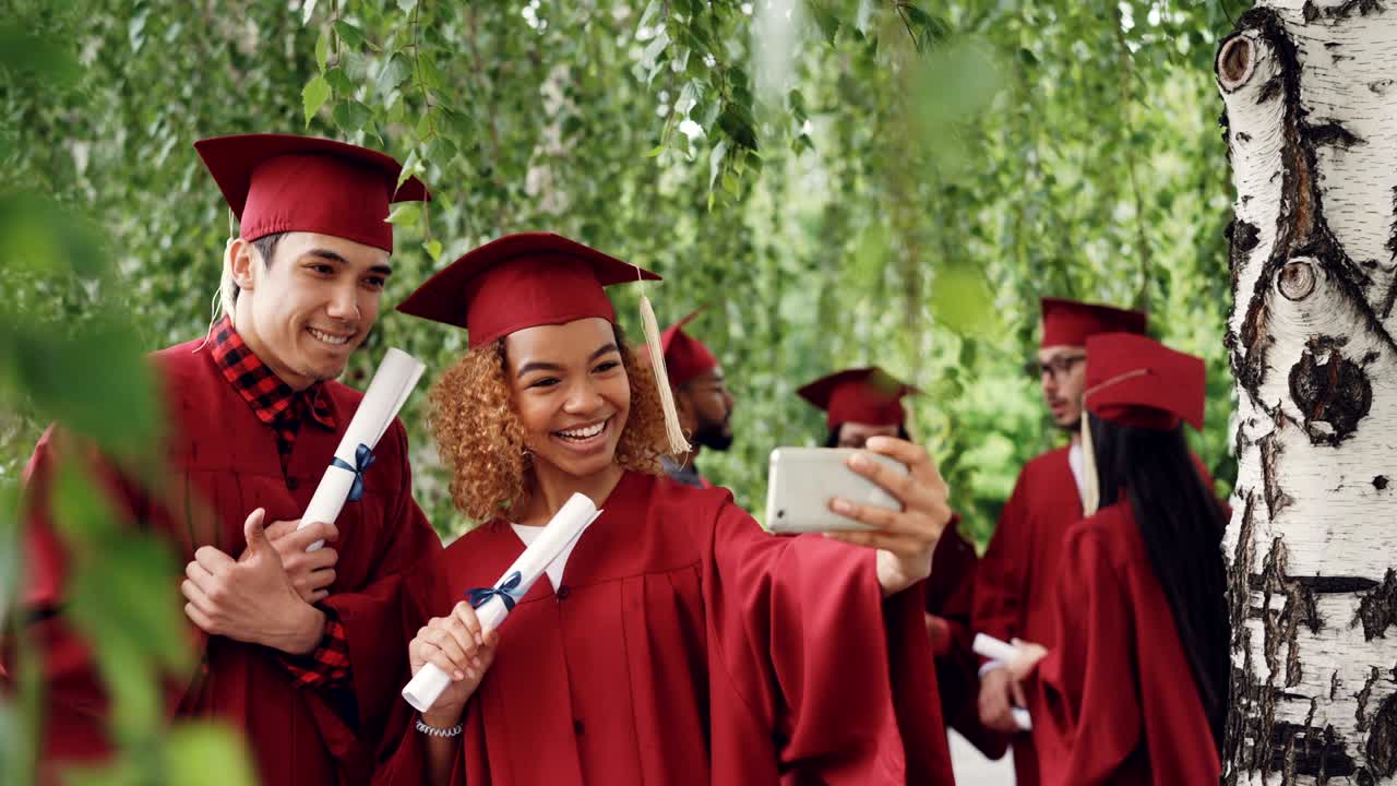 Glade unge mennesker, pige og fyr, tager selfie efter dimissionsceremonien, mens de holder diplomer og er iført kåber og studenterhuer. Koncept for fotografier og uddannelse.