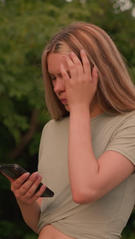 Young woman in cropped top and jeans, focused intently on her phone while walking along a greenery-lined path, with one hand on her ear and the other holding her phone, she appears deep in thought