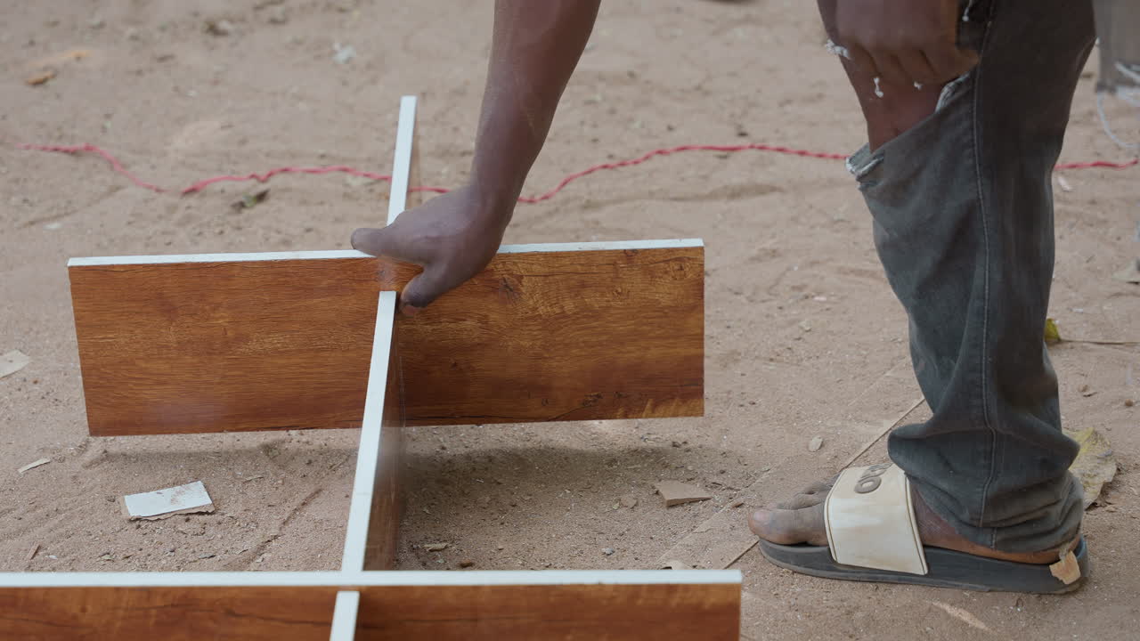 Person assembling wooden divider on sand with closeup of hand guiding melamineedged board into cross joint, torn jeans and sandal visible, outdoor jobsite mood with focused manual alignment for shelf
