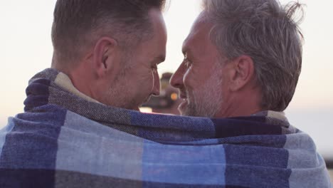 Feliz-Pareja-De-Hombres-Gays-Caucásicos-Vistiendo-Una-Manta,-Abrazándose-Y-Tomándose-Selfie-Al-Atardecer-En-La-Playa