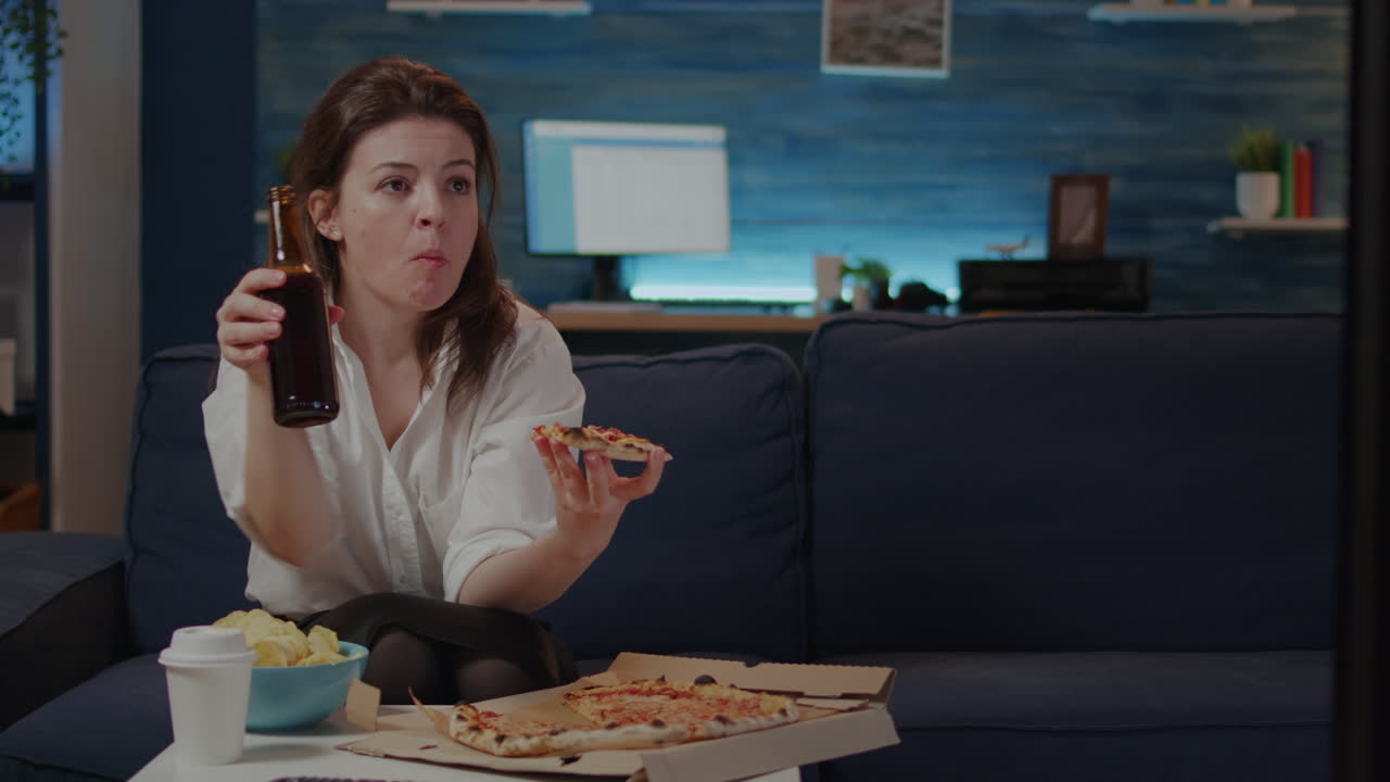Young woman eating slice of pizza from box and drinking beer