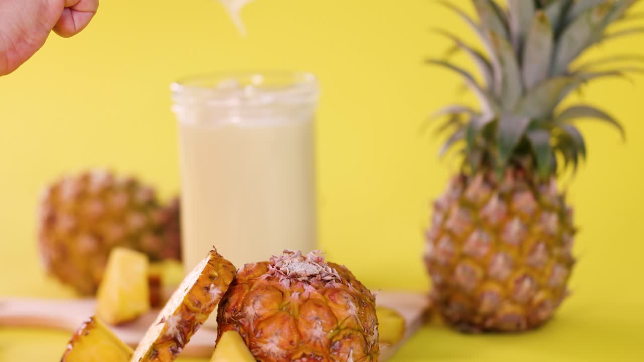 Hand pours creamy pineapple smoothie into glass, surrounded by fresh pineapple slices, bright yellow background
