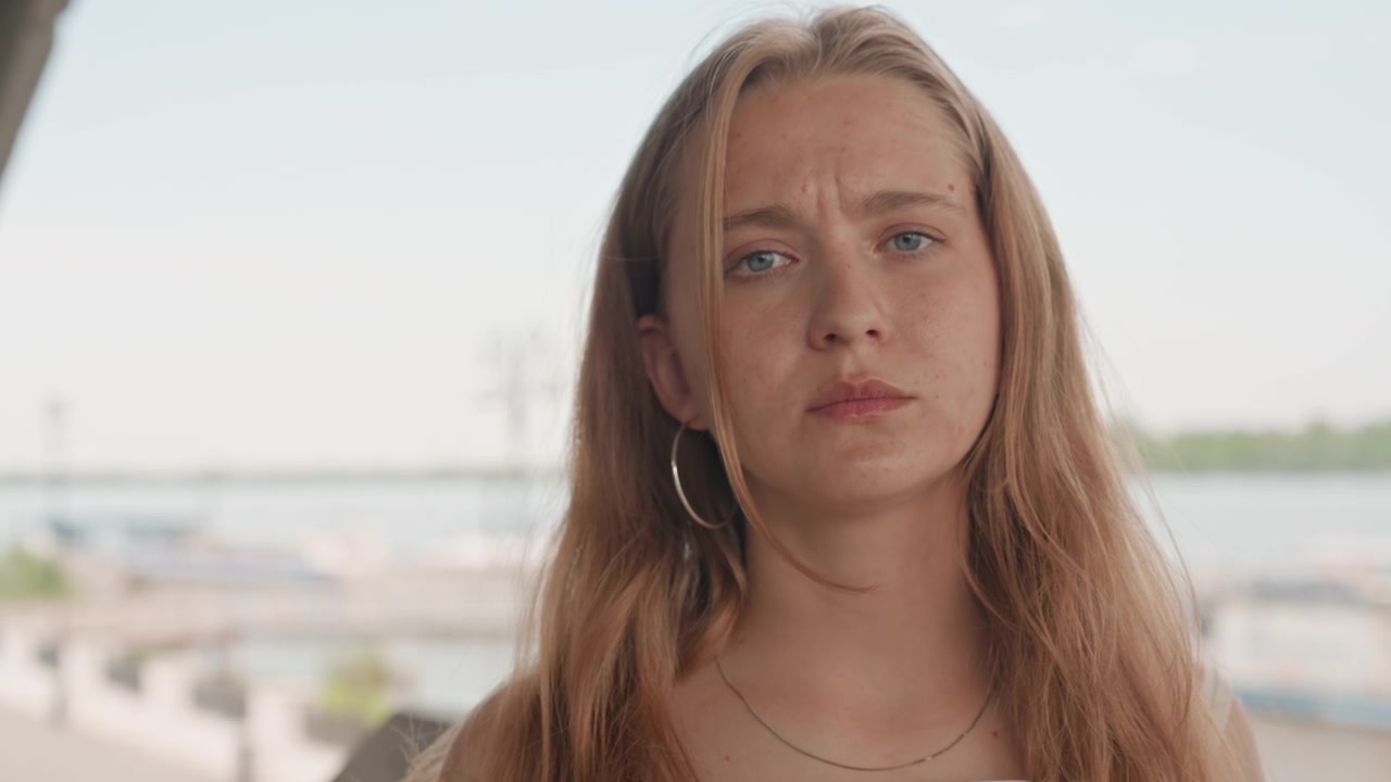 Donna con capelli lunghi e orecchini a cerchio in piedi all'aperto vicino a uno specchio d'acqua, che indossa abiti casual. Lo sfondo mostra una luce diurna soffusa su un paesaggio calmo con strutture distanti