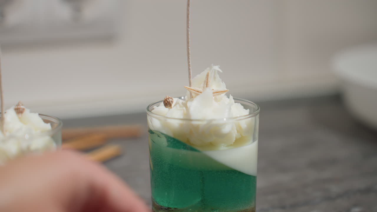 Female decorator places shell ornament delicately on whipped wax topping inside glass cup candle layered with green and white colors, adding final artistic touch to handcrafted home decor piece