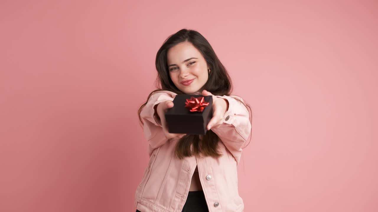Happy woman giving gift box and looking at camera in pink studio