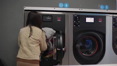 a woman strides toward the machine, loading numerous clothes into the laundromat and readying to activate it