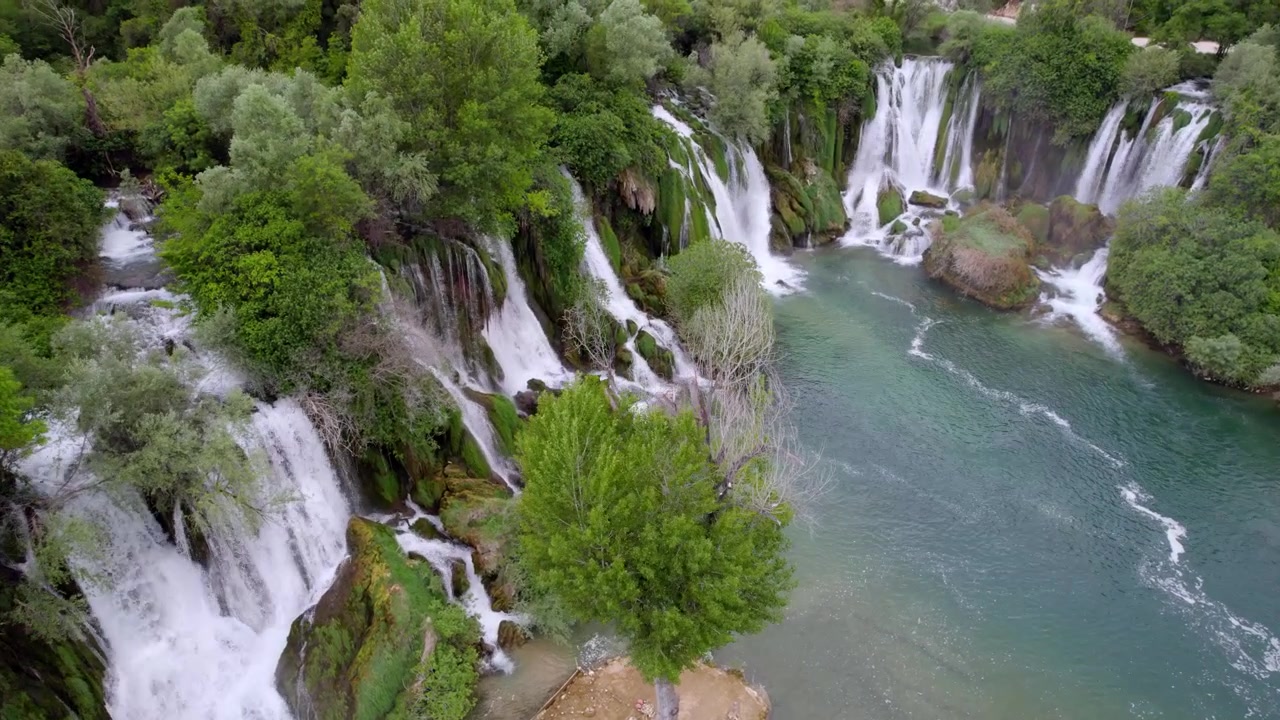 La Cascata di Kravica scorre accanto a un albero solitario in acque limpide e turchesi in Erzegovina, panoramica aerea con dolly di apertura