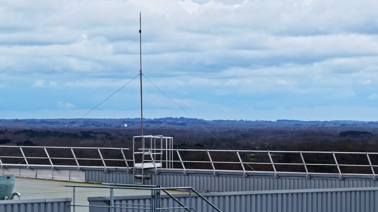 Drone ascending over industrial building, rooftop antenna and solar panels. Rural woodland backdrop. France