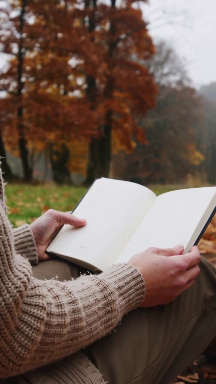 Video verticale: Allontanamento, lettore con maglione color cammello che sfoglia le pagine di un libro tascabile mentre legge al parco.