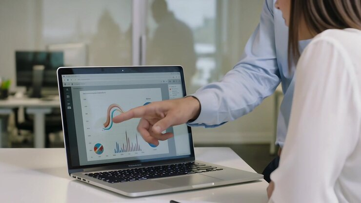 Moving in, man in blue shirt leaning over office table pointing at laptop charts, explaining data