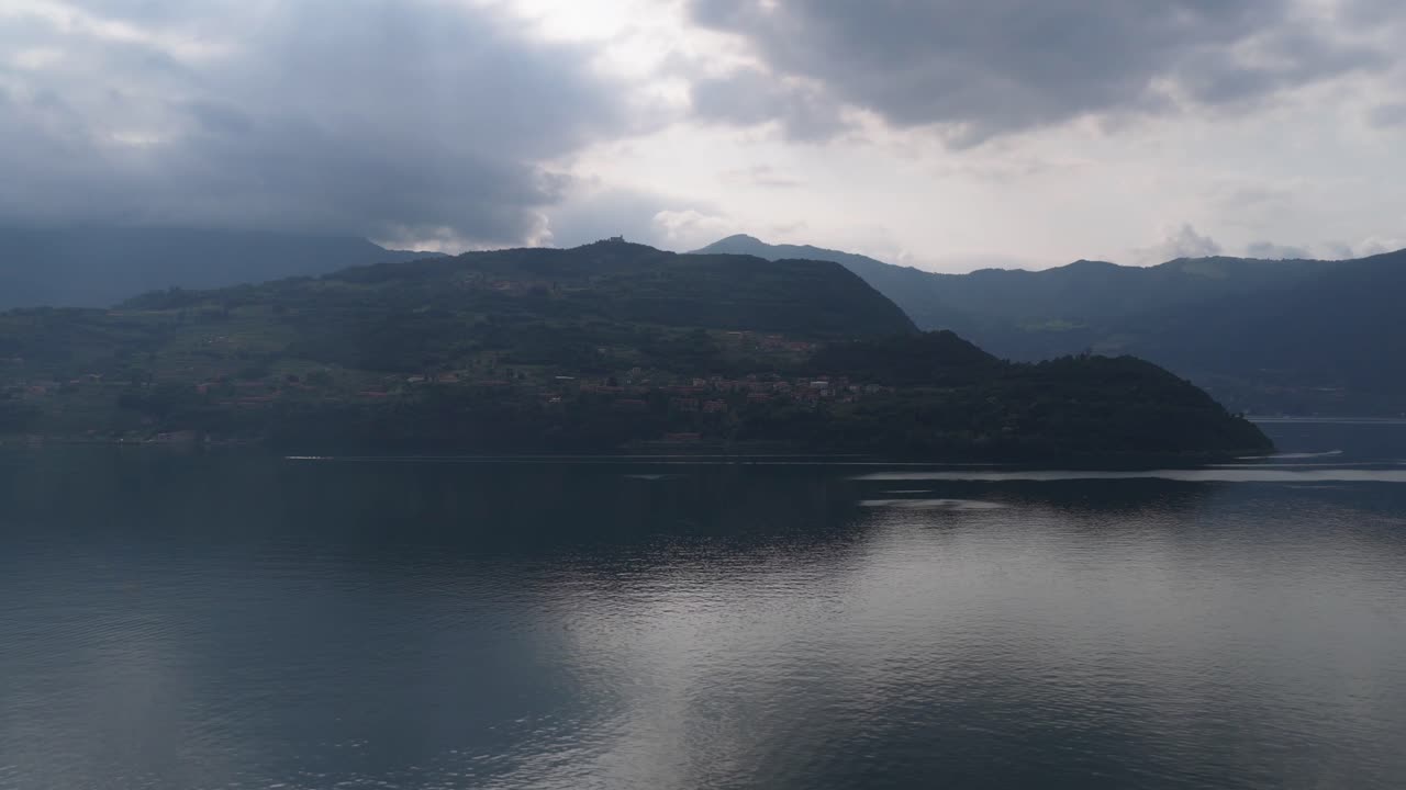 Overcast drone shot of a misty hillside above Lake Iseo with storm clouds reflecting on the lake surface