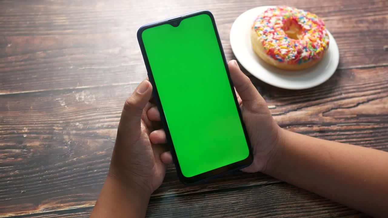 niño sosteniendo un teléfono inteligente con pantalla verde y una rosquilla en una mesa de madera