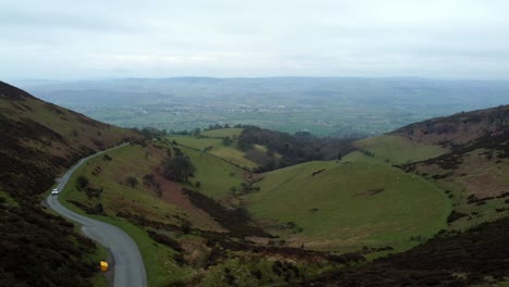 Vehículo-Blanco-Que-Conduce-Un-Camino-Rural-único-Y-Estrecho-Que-Atraviesa-El-Paisaje-De-Valles-De-Montaña-Verde-Galés