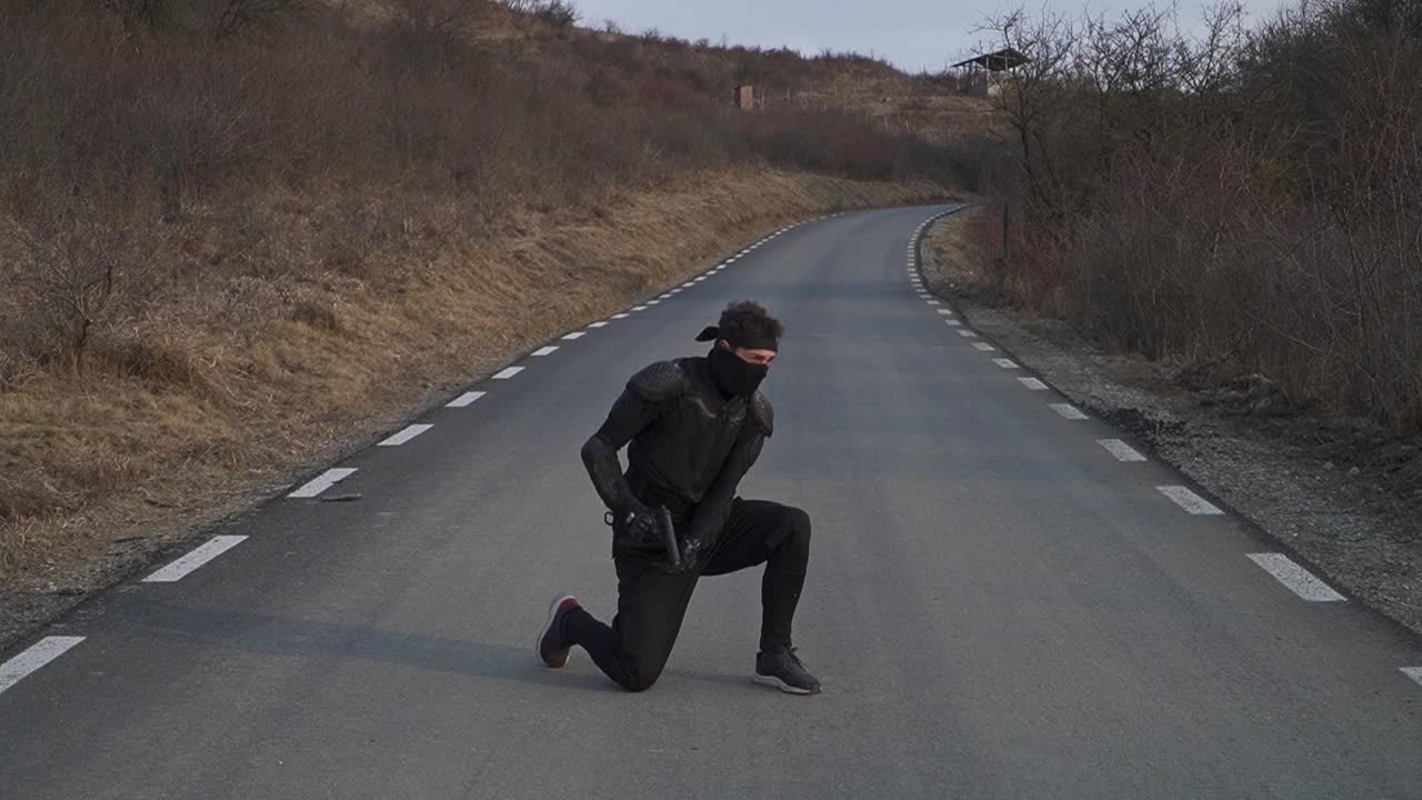 Dynamic action shot of a masked man mid-flip while holding a gun, dressed in black tactical gear on an empty road surrounded by dry, rugged terrain.