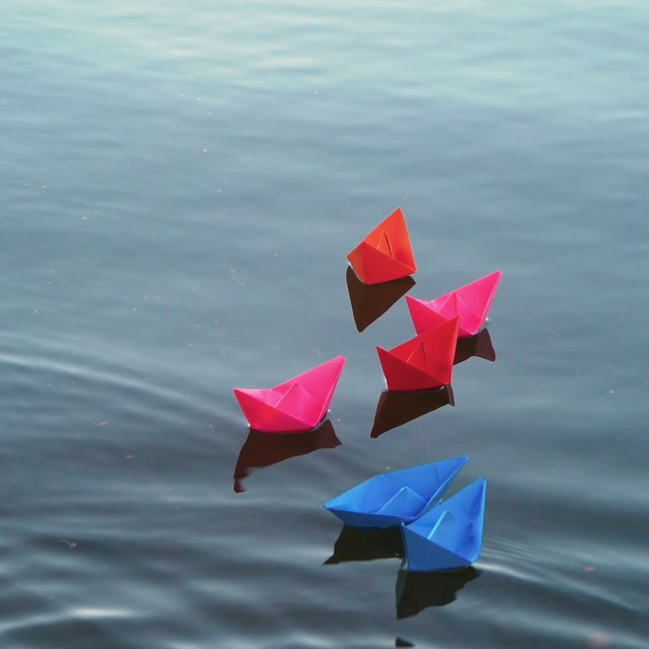 Six colored paper boats on blue water and some light splashes falling on. Beautiful children's origami ships floating in the river. Close-up