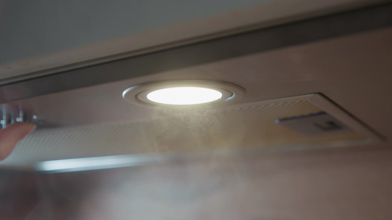 Nærbilde fra lav vinkel viser en kjøkkenvifte i rustfritt stål under et skap, med damp som beveger seg inn i det perforerte filteret, mykt lys fra innfelt belysning, børstet overflate, refleksjoner, dis og et rolig hjem