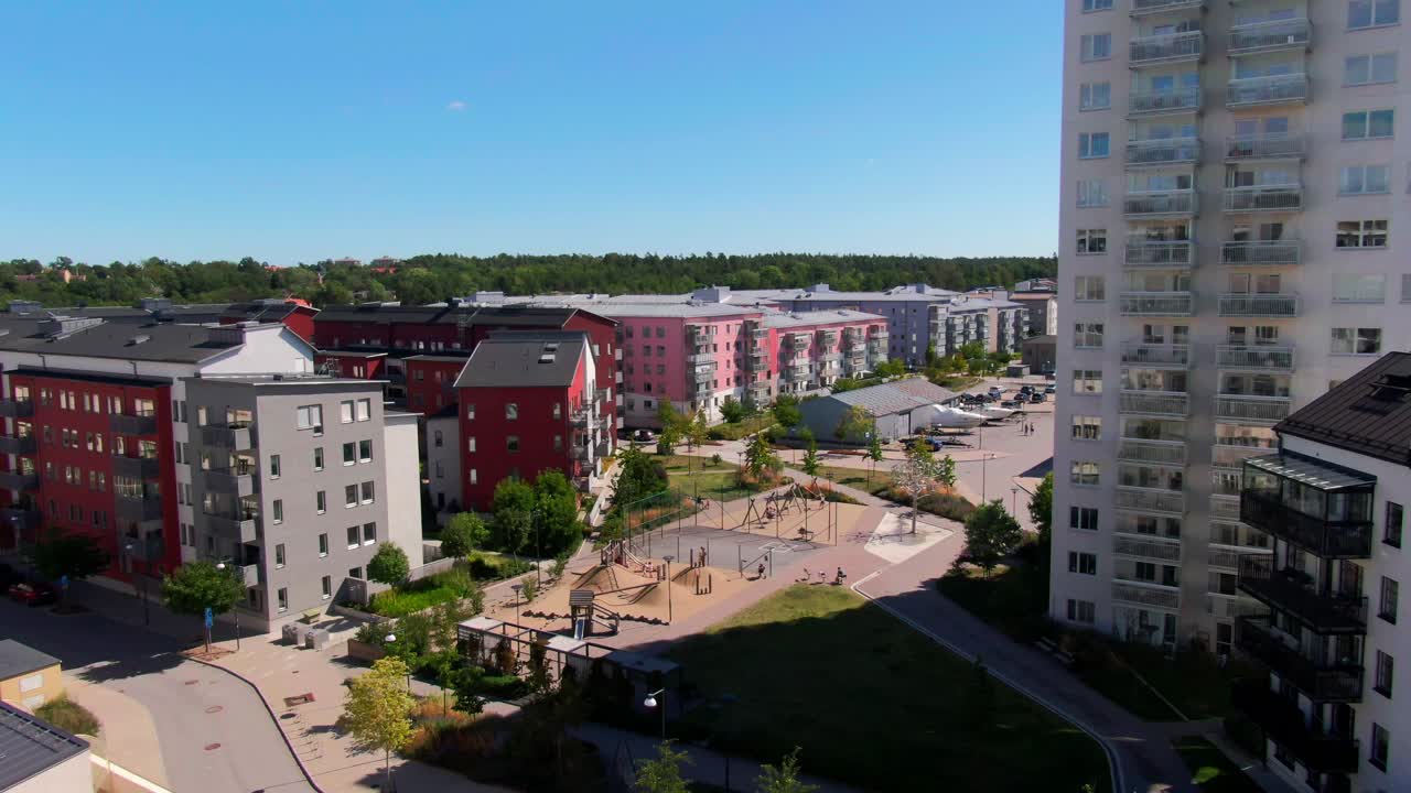 Živý letecký pohled na moderní švédské apartmány s barevnými fasádami, hřištěm a zelenými dvory pod jasnou modrou oblohou v Gustavsberg. Ideální pro stock s rodinným životním stylem