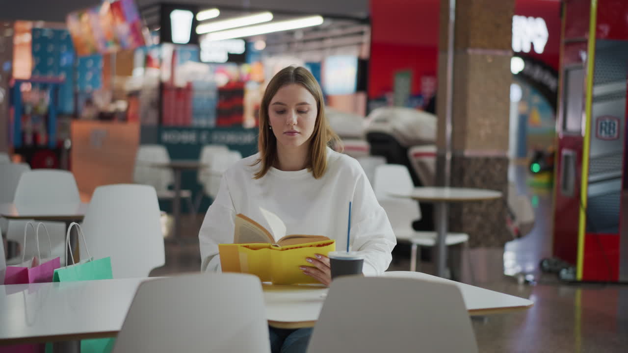 White lady seated in a mall reading a book with a coffee cup and shopping bag nearby, soft lighting creates a relaxed atmosphere with blurred background