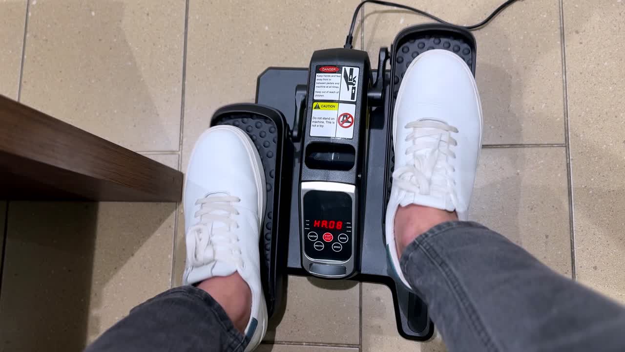 Vista dall'alto in prima persona di una persona con scarpe da ginnastica che si allena su un ellittico compatto da scrivania. Le sue gambe pedalano mentre la console digitale tiene traccia dell'allenamento in ufficio