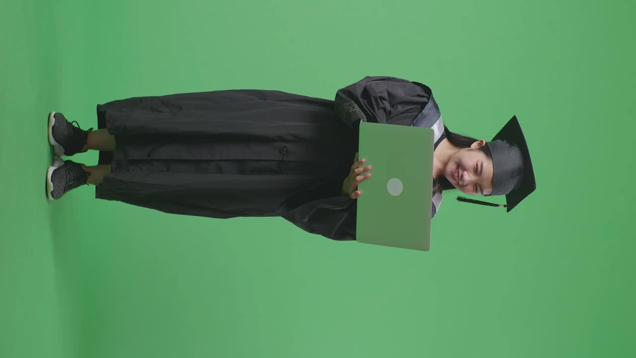 Laureata asiatica a figura intera in tocco e toga che digita su un laptop, in piedi su sfondo green screen in studio