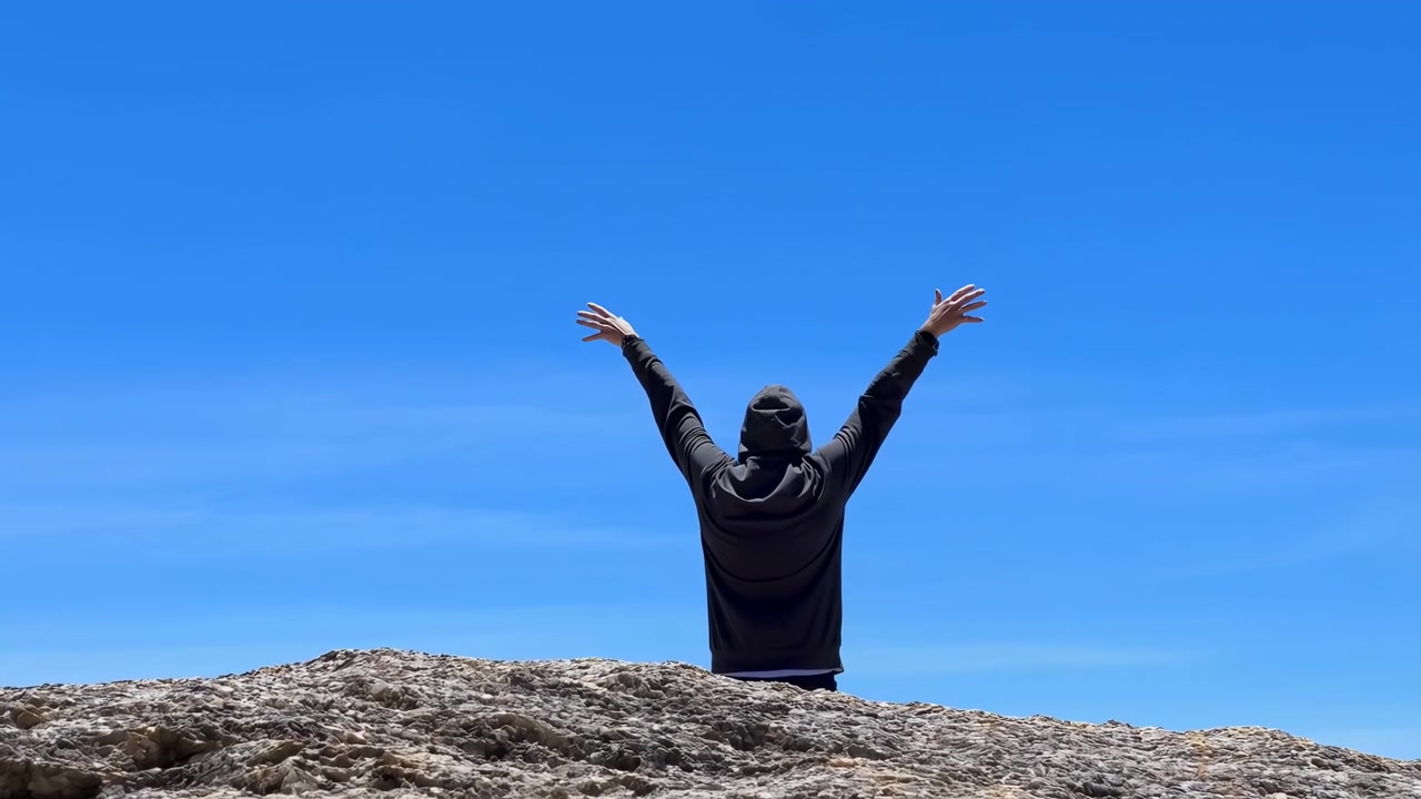 Rear facing person in black hooded sweatshirt stands on textured pale rock ridge, lifting both arms high above head against uninterrupted deep blue sky with calm minimalist landscape and strong