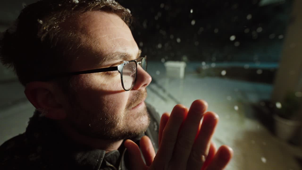 Close-up de um homem em pé ao ar livre à noite enquanto neva forte. Ele assopra as mãos e as esfrega para se manter aquecido. Conceito de clima frio, desconforto no inverno, espera ao ar livre e vulnerabilidade humana