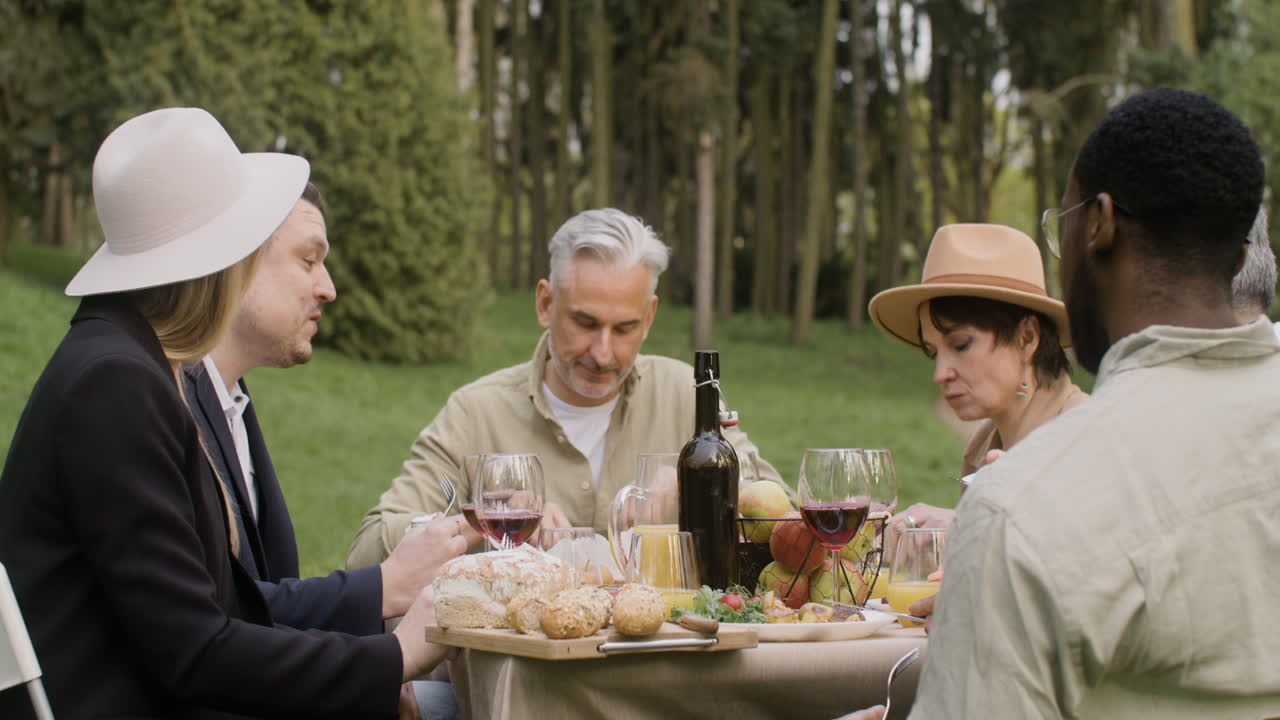 공원에서 야외 파티를 하는 동안 테이블에 앉아 서로 먹고 이야기하는 중년 친구들
