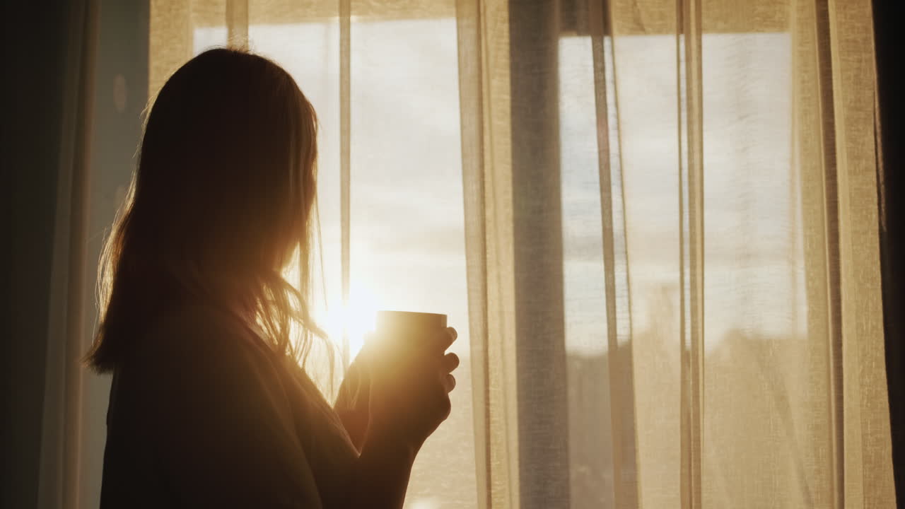 silueta de una mujer con una taza de té, de pie en la ventana al atardecer. vista de atrás
