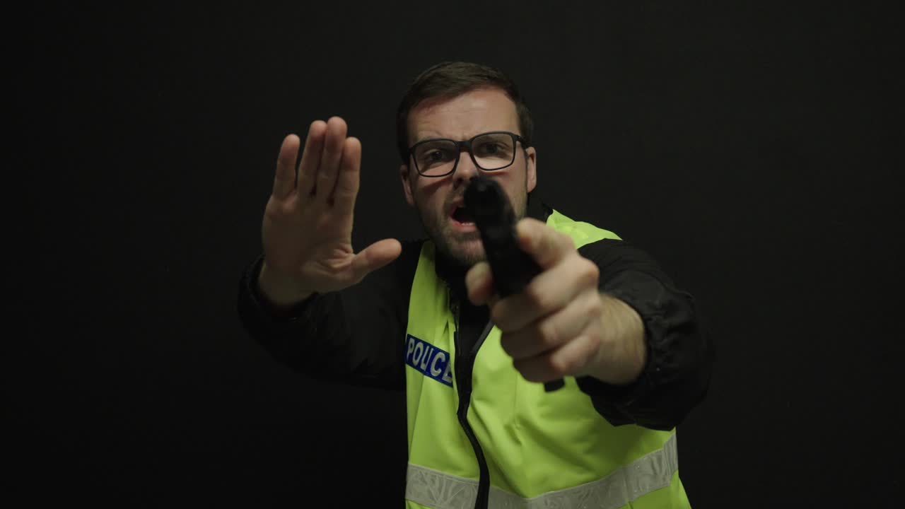 Agente di polizia in gilet di sicurezza che punta un'arma direttamente verso la telecamera su uno sfondo scuro. Concetto di confronto con le forze dell'ordine, risposta alle minacce, autorità e sicurezza pubblica.