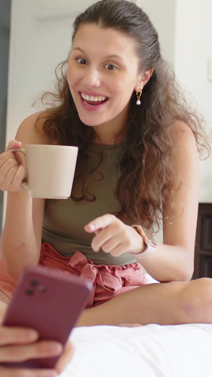 Video vertical: Mujer reaccionando, sonriendo y frunciendo el ceño, sosteniendo una taza en la cama, mientras otra muestra el teléfono y habla.