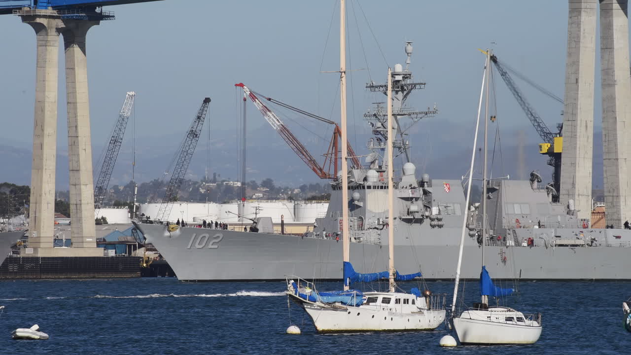 Large naval ship docked near the Coronado Bridge with sailboats and cranes in the harbor