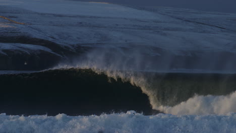 ocean wave crashes as mist rises off top in front of snow covered mountains, nobody riding, slow motion