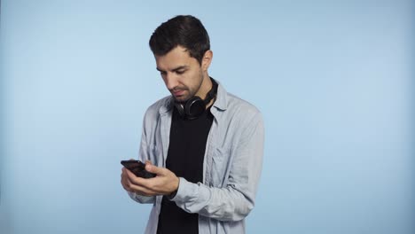 young confident man holding phone and scroll screen or typing message isolated on blue background. wearing headphones on neck