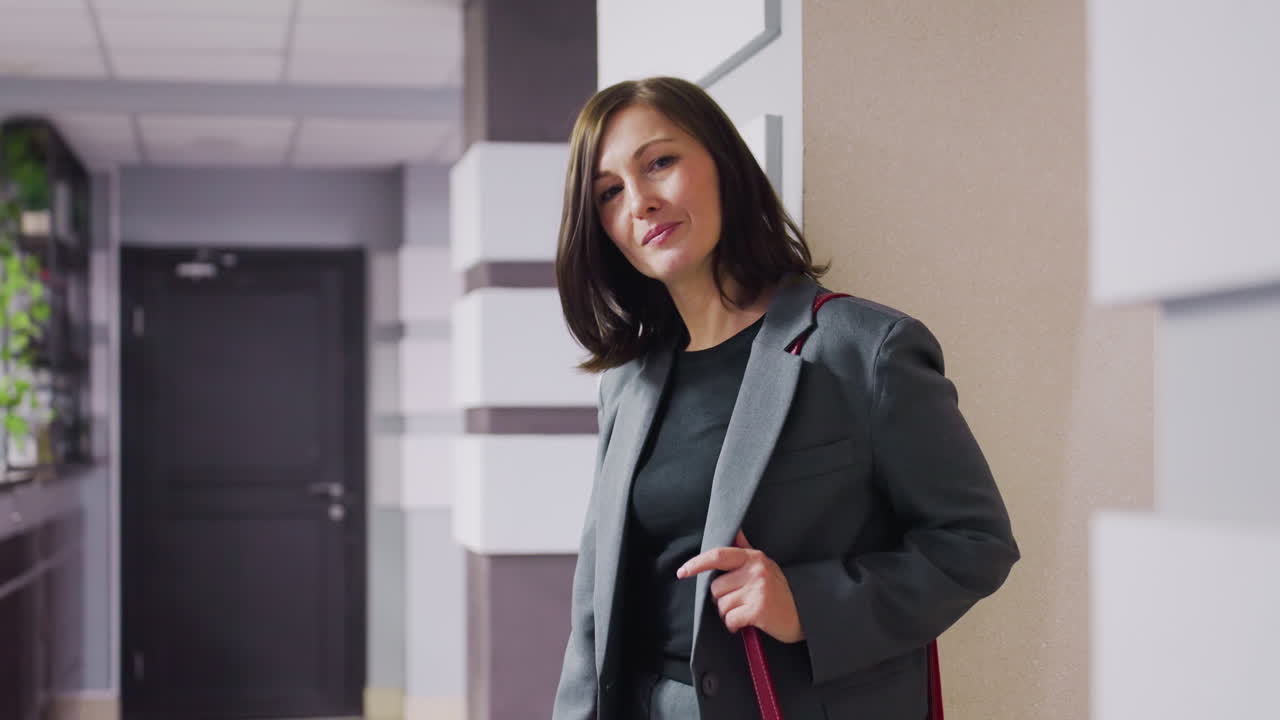 Professional businesswoman in gray suit with red handbag standing confidently in office hallway, looking to side with calm expression, modern office background with sleek design