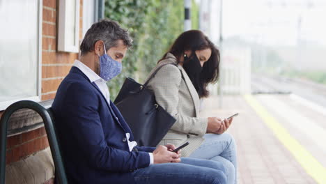 Business-Commuters-On-Railway-Platform-With-Mobile-Phones-Wearing-PPE-Face-Masks-During-Pandemic