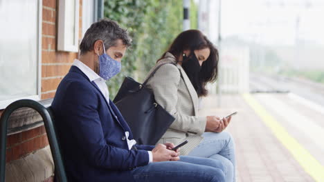 pasajeros de negocios en la plataforma del ferrocarril con teléfonos móviles usando máscaras faciales de ppe durante la pandemia