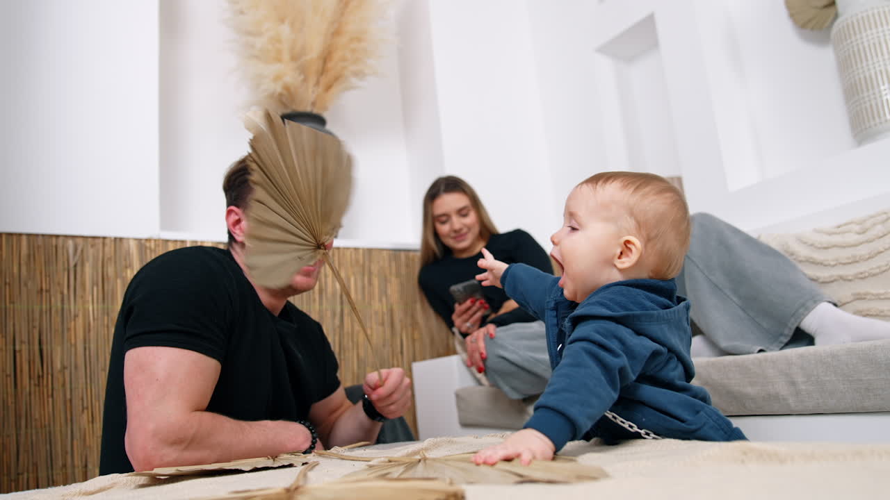 Father is playing with his cute little baby boy. Dad waves a leaf to his son. Mother lies on the sofa holding a phone at backdrop.