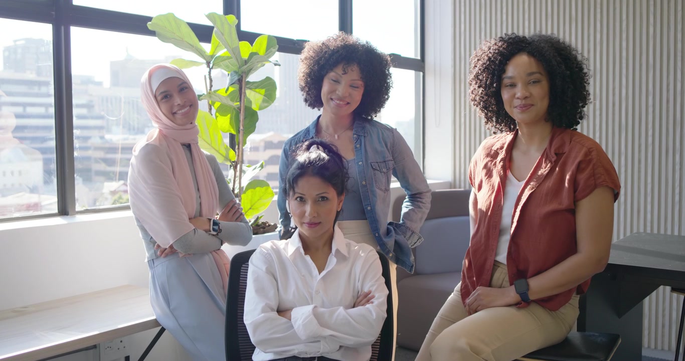 Colegas de trabalho diversas posando para um retrato perto de grandes janelas e uma cadeira ergonômica