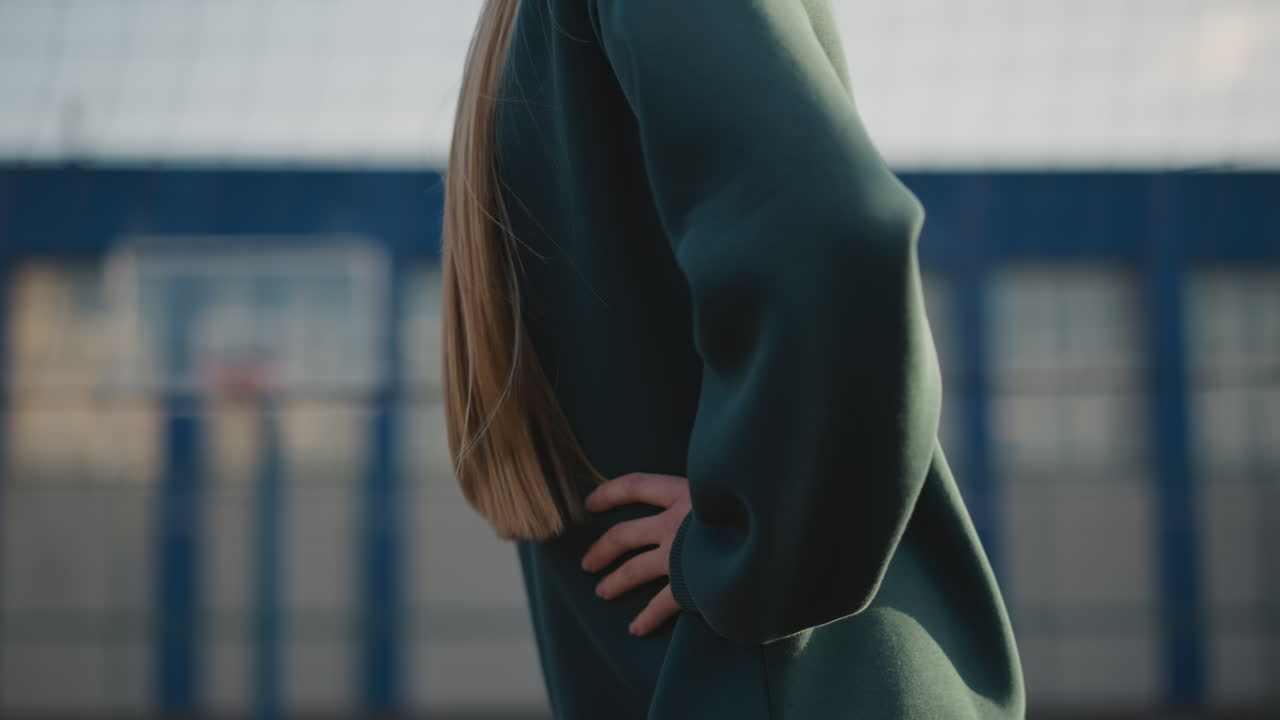 긴 머리카락과 함께 녹색 스웨트 셔츠를 입은 젊은 여자의 클로즈업, 허리 회전 운동, 스포츠 네트와 야외 피트니스 코트 건물을 특징으로하는 흐릿한 배경