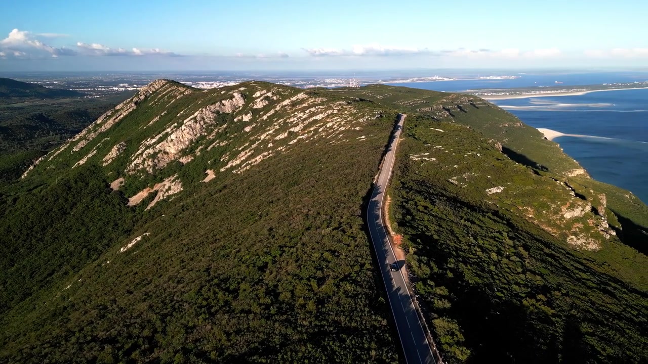 Riprese drone di un'autostrada tortuosa lungo la cresta del Parco Naturale di Arrábida a Setúbal, Portogallo, che mostra rigogliose colline verdi e una costa oceanica distante.