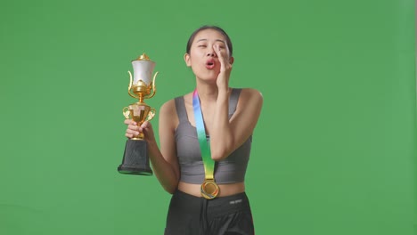 asian woman with a gold medal holding a gold trophy and yelling positively with hand over mouth on green screen background in the studio