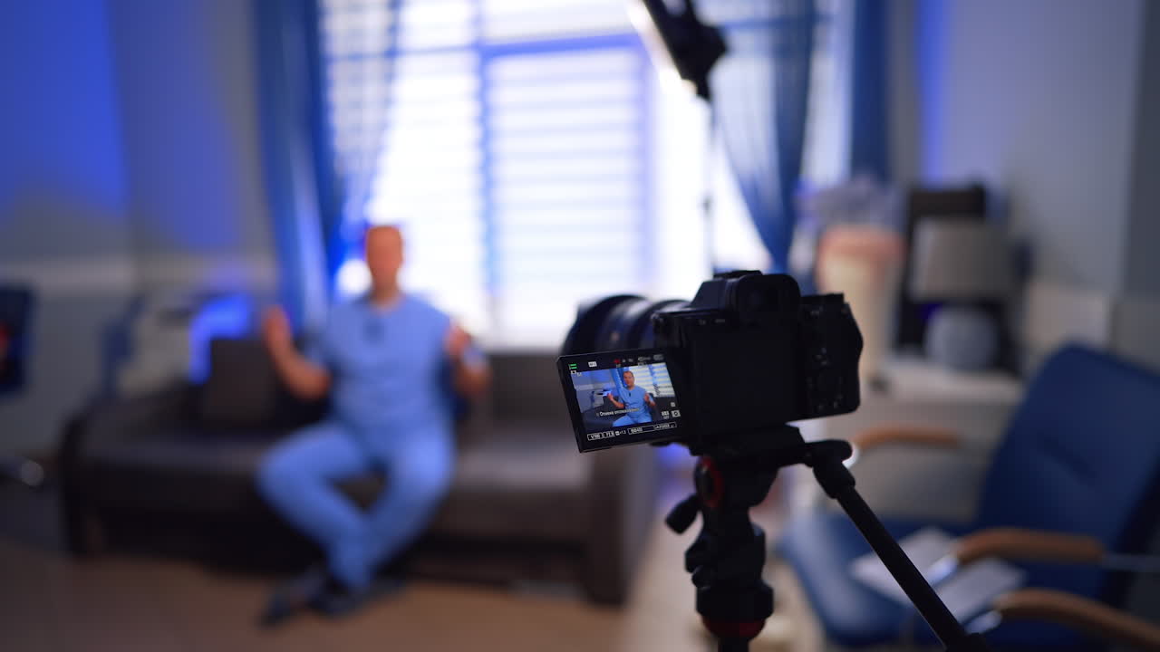 Medical vlog content created indoors. Male wearing blue uniform talks to camera on tripod. Selective focus