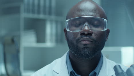 portrait of african american lab scientist in safety glasses with microscope