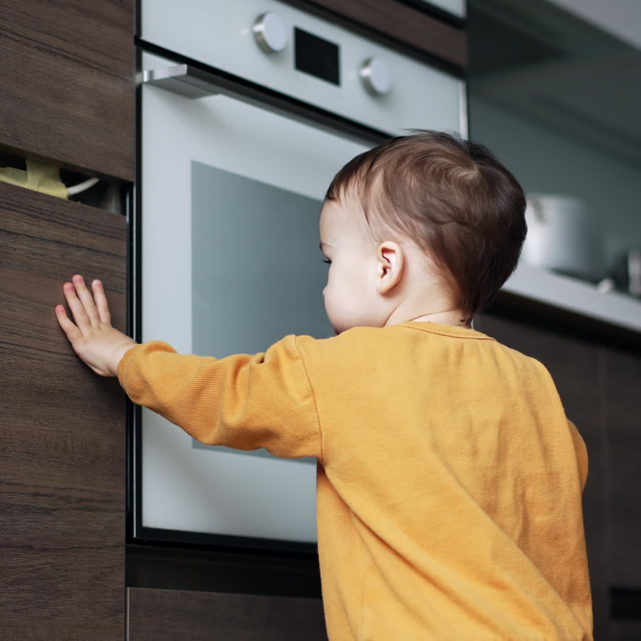 Caucasian toddler in orange suit looks through the kitchen lockers. Little kid opens the door checking what's inside