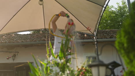 Deslice-La-Toma-En-ángulo-Bajo-De-Una-Mujer-Regando-Las-Plantas-Del-Patio-En-La-Mañana-De-Verano