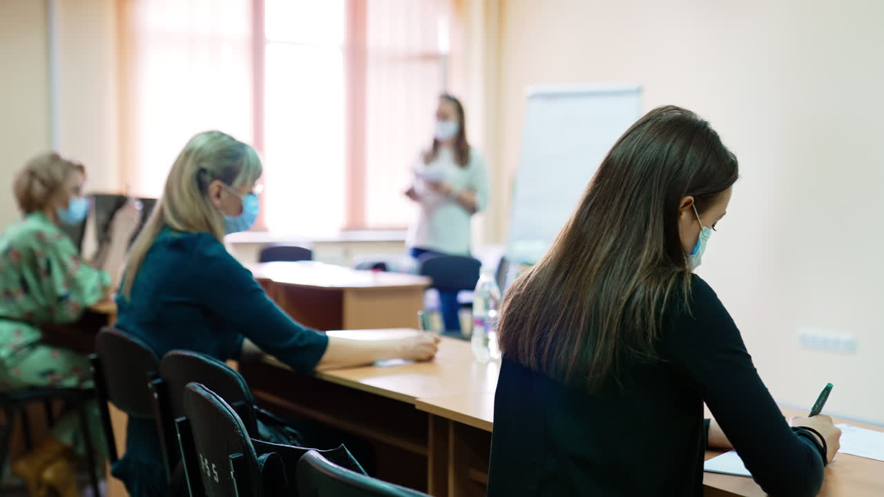 Students wearing surgical masks taking notes of a lecture. Women sit at desks listening to the female lecturer. Blurred backdrop.