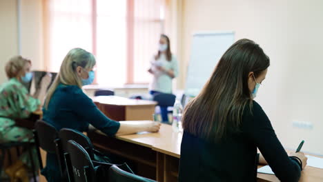 students wearing surgical masks taking notes of a lecture. women sit at desks listening to the female lecturer. blurred backdrop.