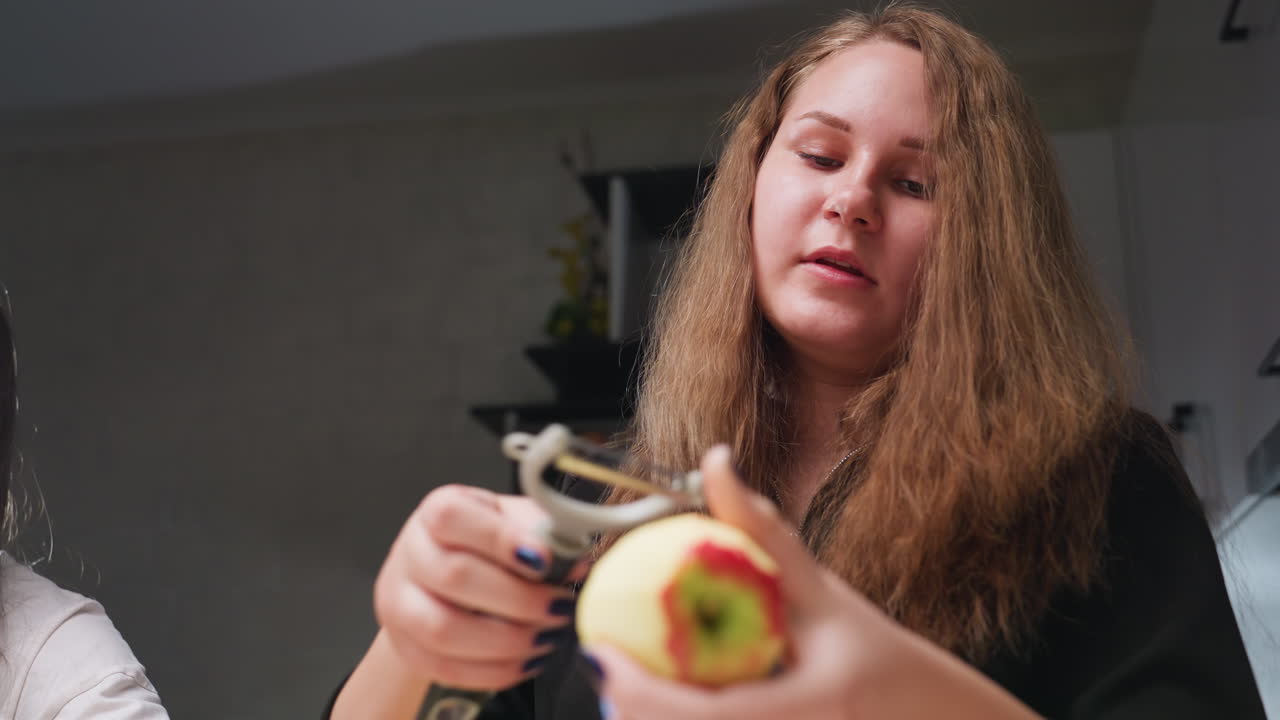Two friends side by side in kitchen converse while one peels apple using peeler, blue manicure visible, soft dim background, candid casual food prep moment, healthy snack vibe, natural light